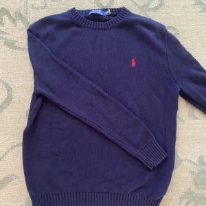 Mörkblå stickad tröja Polo Ralph Lauren - Klassisk mörkblå stickad tröja från Polo Ralph Lauren med rund halsringning och röd broderad logga på bröstet. Tröjan har ribbade muddar vid ärmslut och nederkant, perfekt för lager-på-lager-stil. Pris kan diskuteras 👌
