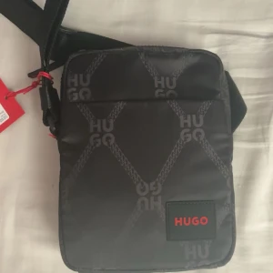 Svart axelväska från Hugo - Snygg svart axelväska från Hugo med logomönster och röd Hugo-detalj framtill. Väskan har justerbar axelrem, dragkedja upptill och är tillverkad i slitstark polyester. Perfekt storlek för mobil, plånbok och andra småsaker.