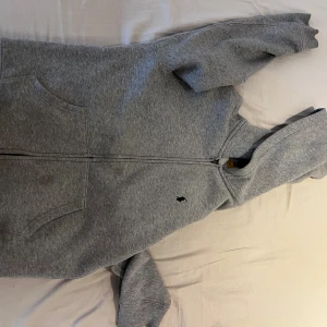 Grå hoodie från Polo Ralph Lauren - Klassisk grå hoodie från Polo Ralph Lauren med dragkedja framtill och två stora fickor. Tröjan har huva, lång ärm och den ikoniska broderade loggan på bröstet. Perfekt för chill dagar och enkel att matcha med jeans eller joggers.