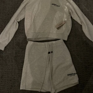 Grå Essentials Fear of God set - Säljer en stilren ljusgrå hoodie från Essentials Fear of God. Tröjan har huva, lång ärm och en klassisk kanguruficka framtill. Diskret logga på bröstet och mjukt material som känns skönt mot huden. Perfekt för chill dagar och streetwear-stil.                 Helt sprillans ny så alltså absolut inga defekter! ❗️nypris 2500kr ❗️