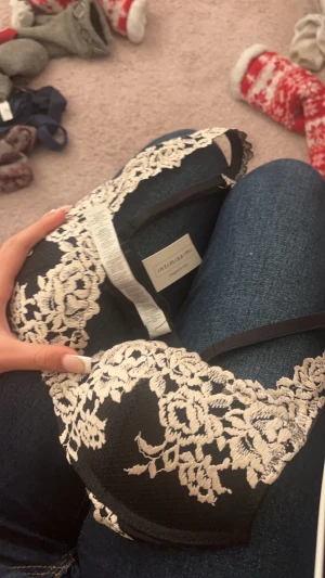 Svart spetsig bandeau från Intimissimi - Supersnygg svart bandeau-topp från Intimissimi med vit spets i blommigt mönster. Toppen har tunna axelband och en elegant, romantisk vibe. Perfekt att styla under en kavaj eller till höga jeans för en trendig look.