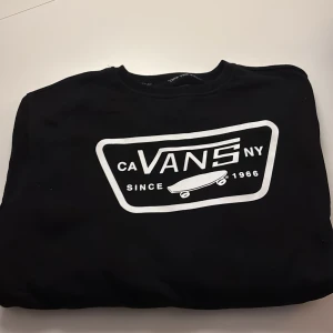 Svart sweatshirt från Vans med tryck - Svart sweatshirt från Vans i storlek small med stort vitt tryck framtill som visar loggan och en skateboard. Rund hals och mjukt material som känns skönt mot huden. Perfekt för dig som gillar streetwear och skatekänsla.
