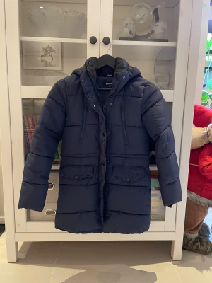 Mörkblå dunjacka från Everest barn strl 146 - Snygg mörkblå dunjacka från Everest med huva, långa ärmar och flera praktiska fickor med både dragkedja och knappar. Jackan har quiltad design och reflekterande detalj baktill. Perfekt för kalla dagar och har en längre passform. Storlek 146