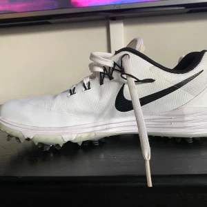 Vita golfskor från Nike med dobbar - Använda en golfrunda. Snygga vita golfskor från Nike med svarta detaljer och stor svart swoosh på sidan. Skorna har snörning, vadderad häl och slitstark syntetisk ovandel. Yttersulan har flera dobbar för extra grepp på golfbanan. Mellansulan är märkt med Lunarlon för extra komfort.