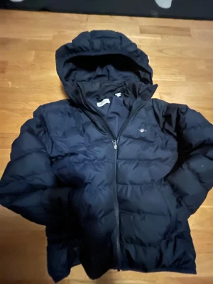 Mörkblå pufferjacka från GANT - Säljer en mörkblå pufferjacka från GANT med huva och dragkedja. Jackan har GANT-logga med sköld på bröstet och är vadderad för extra värme. Perfekt för kalla dagar och har en klassisk, sportig look. Inga fläckar eller några problem under min tid funkar perfekt 