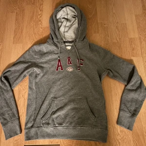 Grå hoodie Abercrombie & Fitch - Snygg grå hoodie från Abercrombie & Fitch med broderad A&F-logga i vinrött framtill. Klassisk känguruficka, dragsko i huvan och mjukt bomullsmaterial. Perfekt för en chill och avslappnad stil.