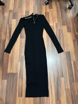 Svart långärmad klänning Marciano - Snygg svart ribbad långklänning från Marciano Los Angeles med lång ärm och cool asymmetrisk axel. Klänningen har dekorativa guldknappar och dragkedja vid halsen för extra edge. Figurnära passform som ger en stilren siluett.