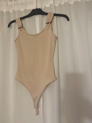 Beige ribbad body med ringdetaljej - Säljer en beige ribbad bodysuit med breda axelband och snygga ringdetaljer i metall vid axlarna. Använd den 1 gång är som ny