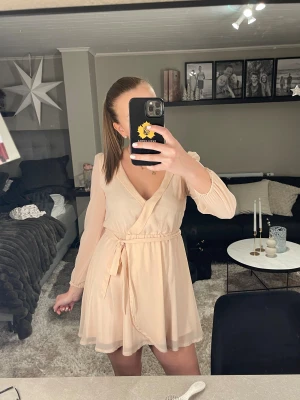 Ljus omlottklänning från NLY Trend - Supersöt ljusbeige omlottklänning från NLY Trend med v-ringning och långa, transparenta ärmar. Klänningen har knyt i midjan och ett flowy, kort fall. Perfekt för dig som gillar en feminin och enkel stil.