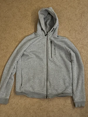 Grå hoodie från Sail Racing - Snygg grå hoodie från Sail Racing med dragkedja hela vägen, huva med snörning och en bröstficka med dragkedja. Mjuk insida och ribbade muddar. Perfekt för chill dagar eller när du vill ha en sportig look.