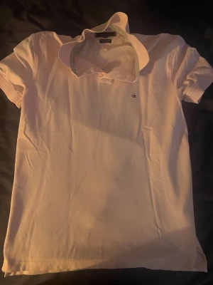 Ljusrosa pikétröja Tommy Hilfiger - Klassisk vit pikétröja från Tommy Hilfiger, modell The 1985 Polo Shirt. Tröjan har korta ärmar, krage med diskreta ränder på insidan och liten broderad logga på bröstet. Tillverkad i bomull för en skön och luftig känsla.