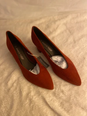 Rostbruna pumps från Marco Tozzi - Snygga rostbruna pumps från Marco Tozzi med spetsig tå och låg klack. Skorna är tillverkade i mjuk mocka och har en klassisk, stilren form som ger en cool och elegant vibe. Perfekt för dig som gillar att sticka ut med färg och form.