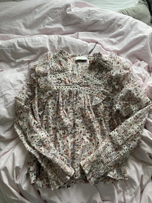 Blommig volangblus från Neo Noir - Romantisk blus från Neo Noir med blommigt mönster i lila, rosa och beige toner. Blusen har volangdetaljer, smockade ärmslut och dekorativa spetsband. Lätt och luftig bomullskvalitet, perfekt för en trendig look.