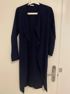 Mörkblå trenchcoat med bälte från C - Snygg mörkblå trenchcoat från C i storlek XS. Jackan har lång ärm, omlott-design och ett matchande bälte i midjan. Stilren och enkel look med mjukt tyg och klassisk krage. Perfekt för dig som gillar minimalistisk stil och vill ha en jacka som funkar till många outfits.