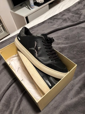 Axel Arigato svarta sneakers med fågel - Svarta sneakers från Axel Arigato med broderad fågel på sidan och vit platt sula. Skorna har snörning, rund tå och är tillverkade i skinn med detaljer i mocka. Snygg minimalistisk design med texten '140MM' på hälen. Kommer med originalkartong och dustbag.