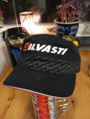 Ny Svart Silvasti keps  - Svart keps från Silvasti med vit och orange broderad logga framtill och på sidan. Kepsen har ett mönstrat band med upprepade Silvasti-tryck runt om och justerbar rem baktill. Materialet är slitstark bomull och skärmen har en röd detalj undertill.
