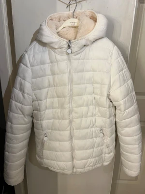 Vit vändbar teddy- och pufferjacka - Supersnygg vändbar jacka i storlek.     S/M. Ena sidan är quiltad puffer i vit polyester med dragkedja och fickor, andra sidan är mjuk teddy i ljusbeige pälsimitation. Jackan har huva och lång ärm, perfekt för kalla dagar. Stilren och mysig look.
