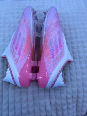 Adidas F50 rosa fotbollsskor - Säljer ett par Adidas F50 fotbollsskor i en snygg mix av rosa och vitt. Skorna har en strömlinjeformad design med platt sula och mönstrade detaljer på sidan. Tillverkade i syntetmaterial för lätt känsla och bra grepp på planen. Perfekt för dig som vill sticka ut på matchen.