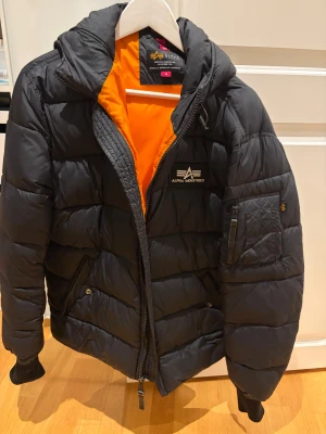 Svart pufferjacka Alpha Industries - Snygg svart pufferjacka från Alpha Industries med orange foder, huva och flera fickor. Jackan har en tydlig logga på bröstet, dragkedja framtill och ribbade muddar vid ärmslut. Perfekt för kalla dagar och har en cool streetstil.