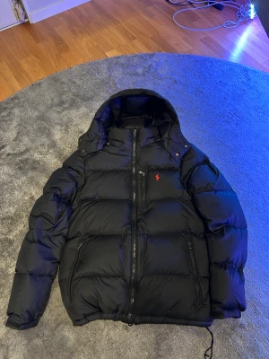 Svart pufferjacka från Polo Ralph Lauren - Säljer en svart pufferjacka från Polo Ralph Lauren med huva som går att ta av. Jackan har en riktigt schysst, puffig look. Perfekt för kalla dagar. Den har används några fåtal gånger förra vintern, otroligt sparsamt använd. Pris kan diskuteras. 