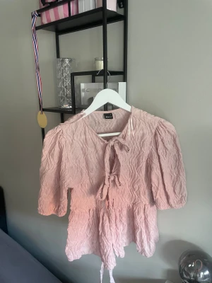 Ljusrosa blus med puffärm  - Supersöt ljusrosa blus  med puffiga trekvartsärmar och crinkle-struktur. Blusen har volang nedtill och knytning fram vid halsen. Materialet är lätt och ger en cool textur. Perfekt för dig som gillar romantisk och trendig stil.