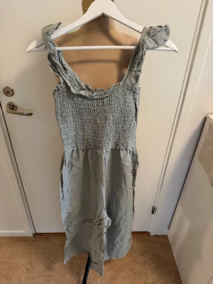Ljusgrön smockad jumbsuit från Asos - Supersöt ljusgrön jumpsuit från Asos med smockad överdel och breda axelband med twistad detalj. Jumbsuit är ärmlös och har en luftig, rak passform som slutar strax under knäet. Perfekt för varma dagar och enkel att styla med sneakers eller sandaler.