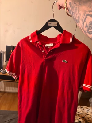 Röd pikétröja från Lacoste - Snygg röd pikétröja från Lacoste med klassisk krage och vita ränder på ärmslut och krage. Tröjan har korta ärmar och den ikoniska krokodilloggan på bröstet. Perfekt för dig som gillar sportig och stilren look.