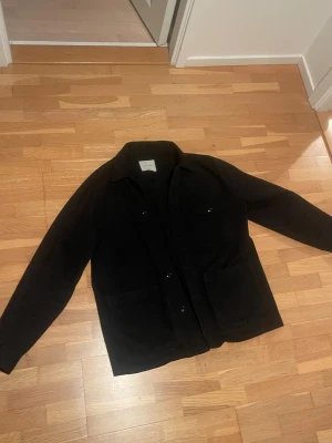 Svart overshirt från Arket - Svart/mörkblå overshirt från Arket med klassisk krage, tre stora fickor framtill och knäppning med svarta knappar. Jackan är tillverkad i mjuk bomull och har en clean, stilren look som funkar till många outfits. Perfekt för lager-på-lager under hösten.