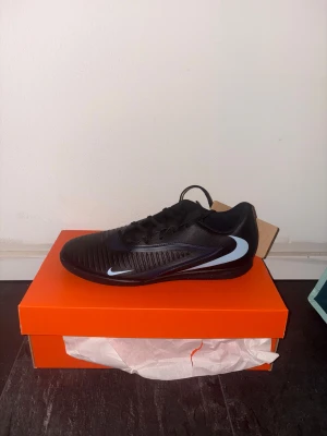 Nike Phantom 6 Inomhusskor - Snygga svarta Nika phantom 6. Skorna har snörning och en platt sula med mönster för bra grepp på inomhusplan. Ovandelen är tillverkad i syntetmaterial med diskreta ribbade detaljer för bättre bollkontroll. 