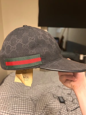 Svart Gucci keps med GG-mönster  - Snygg svart keps från Gucci med diskret GG-mönster över hela kepsen och klassiska gröna och röda ränder på sidan. Kepsen har böjd skärm och är tillverkad i bomull. Perfekt för dig som vill ha en lyxig streetstyle-look. 