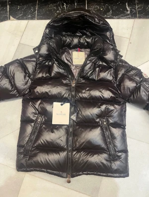 Svart Moncler dunjacka med huva - Snygg svart dunjacka från Moncler med glansig finish och huva. Jackan har två dragkedjeförsedda fickor framtill, quiltad design och klassisk Moncler-logga på ärmen. Perfekt för kalla dagar och riktigt stilren look.