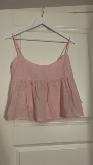 Rosa linnetopp med rosett från Nelly - Supersöt ljusrosa linnetopp från Nelly med tunna axelband och volang nedtill. Toppen har en snygg rosett i ryggen och är tillverkad i ett luftigt linnematerial med diskret rutmönster. Perfekt för varma dagar och chill vibes.