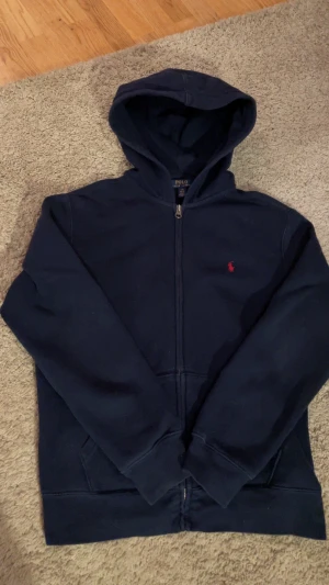 Marinblå hoodie från Polo Ralph Lauren - Snygg marinblå hoodie från Polo Ralph Lauren med dragkedja och huva. Klassisk röd logobrodering på bröstet och två fickor framtill. Perfekt för chill dagar och enkel att matcha med jeans eller joggers.