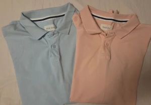 Pier One ljusblå och ljusrosa polotoppar - Två stilrena polotoppar från Pier One, en i ljusblå och en i ljusrosa. Båda har klassisk krage, korta ärmar och knappslå framtill. Materialet är mjuk bomull som känns skönt mot huden. Perfekt för dig som gillar enkel och fräsch stil.