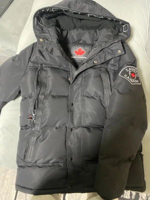 Svart pufferjacka från Zavetti Canada - Svart pufferjacka från Zavetti Canada med broderad logga på ärmen och stor huva med textdetalj. Jackan har två vertikala dragkedjefickor fram, tryckknappar och quiltad design. Perfekt för kalla dagar med sin tjocka och isolerande polyesterfyllning.