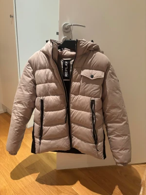 Beige pufferjacka från Replay - Säljer en beige pufferjacka från Replay med huva och svarta detaljer. Jackan har quiltad design, dragkedjor på fickorna och en snygg Replay-logga på ärmen. Perfekt för dig som vill ha en stilren och varm jacka med cool streetkänsla.