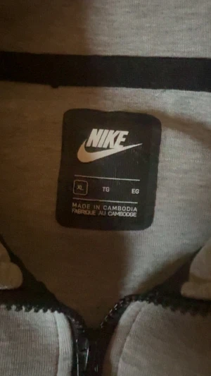 Grå Nike Tech Fleece hoodie XL - Snygg grå Nike Tech Fleece hoodie i storlek XL med svarta detaljer och dragkedja hela vägen. Har huva, två fickor fram och en extra ficka med dragkedja på ärmen. Perfekt för dig som gillar sportig och clean stil.
