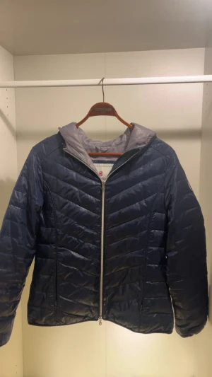 Mörkblå pufferjacka från Conte of Florence - Snygg mörkblå pufferjacka från Conte of Florence Sport med quiltad design och huva. Jackan har dragkedja framtill och ljusgrått foder. Perfekt för kyliga dagar när du vill hålla stilen och värmen.