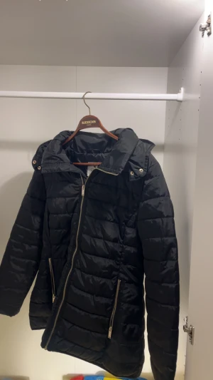 Svart pufferjacka från Rosebud med huva - Svart pufferjacka från Rosebud i storlek M. Jackan har en stor huva, två sidofickor med dragkedja och en grov guldfärgad dragkedja framtill. Quiltad design och snygg, lite längre passform som håller dig varm under kalla dagar.