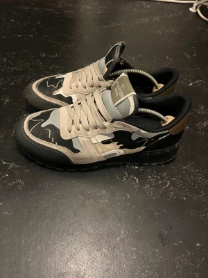 Valentino Garavani Rockrunner sneakers - Snygga Valentino Garavani Rockrunner sneakers med grå, beige och svarta camouflagemönster. Skorna har platt sula, rund tå och detaljer i mocka och skinn. Klassisk snörning och coola rockstuds på hälen. Kommer med originalkartong och tags. Väldigt bra skick , hör av er vid frågor!