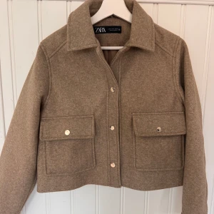 Beige overshirt från Zara i ullmix - Stilren beige overshirt från Zara i ullmix med klassisk krage, stora fickor framtill och snygga guldfärgade knappar. Jackan har en boxig, croppad passform och är perfekt för lager-på-lager. Enkel att styla till både jeans och kjol.