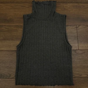 Mörkgrå ribbad polotopp ärmlös - Säljer en mörkgrå ärmlös polotopp med ribbad struktur och hög krage. Toppen har en mjuk och stretchig känsla, perfekt för att styla lager på lager. Avslutet nertill har en lätt vågig kant för extra detalj.