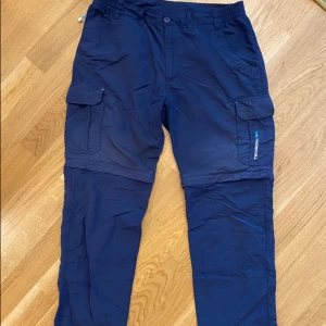 Blå cargopants från Tenson med zip-off - Mörkblå cargopants från Tenson med avtagbara ben så de kan bli shorts. Byxorna har flera fickor med lock och dragkedja vid knäna för enkel omvandling. Perfekta för uteliv och äventyr, tillverkade i lätt syntetmaterial. Storlek M/ 40