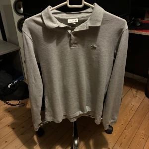 pikétröja Lacoste grå - Snygg grå långärmad pikétröja från Lacoste med klassisk krage och knäppning framtill. Ikoniska krokodilloggan på bröstet. Tillverkad i 100% bomull för en skön och avslappnad känsla. Perfekt för dig som gillar stilren och enkel design.