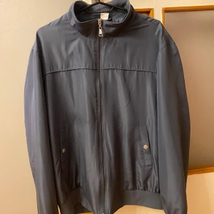 Grå vindjacka med dragkedja - Snygg grå vindjacka i storlek M med hög krage och hel dragkedja framtill. Jackan har två fickor med tryckknappar samt två inne fickor och ribbade muddar vid nederkant och ärmslut. Perfekt för dig som gillar stilren och enkel design.