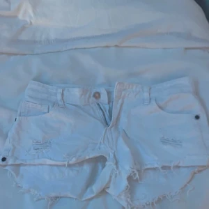 Vita jeansshorts med fransig kant - Snygga vita jeansshorts med fransig och ruffig kant nertill. Klassisk femficksmodell med knapp och dragkedja framtill samt slitna detaljer på framsidan för en cool look. Perfekta för varma dagar.