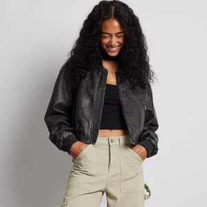 Svart croppad skinn bomberjacka - Säljer en svart croppad bomberjacka i skinnimitation med dragkedja framtill och ribbade muddar. Jackan har en klassisk bomberpassform och är perfekt för dig som vill ha en edgy och stilren look. Enkel att matcha med både jeans och kjol. CROPPAD sitter som bilden i storleken 