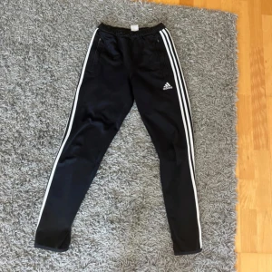 Svarta Adidas träningsbyxor strl 152 - Svarta träningsbyxor från Adidas med klassiska vita ränder längs sidorna och Adidas-logga på låret. Byxorna har resår i midjan, dragkedjor vid fickorna och är tillverkade i ett glansigt syntetmaterial. Perfekta för sport eller chill.