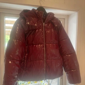 Vinröd pufferjacka från Nelly - Säljer en glansig vinröd pufferjacka från Nelly med huva och dragkedja framtill. Jackan är quiltad och har en oversized vibe, perfekt för kalla dagar. Materialet är syntet och jackan har en riktigt snygg, modern look. Nypris: 799