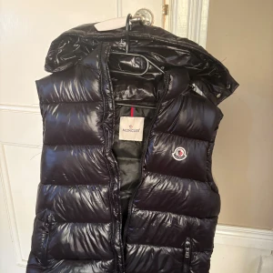 Moncler Väst storlek M/2 - Säljer en svart dunväst från Moncler med glansig finish och avtagbar huva. Västen har dragkedja framtill, två fickor med dragkedja och Moncler-logga på bröstet. Insidan har en cool serietidningsdetalj. Perfekt för dig som vill ha en snygg och varm look.
